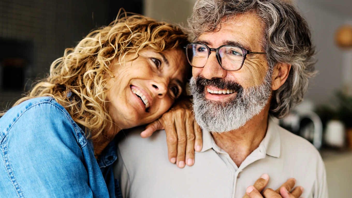 Smiling middle-aged couple at home, the curly-haired woman resting her head and hand on her bearded husband’s shoulder as they look off-camera.