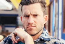 Man with short brown hair and stubble frowning skeptically at the camera with his hand at his chin, seated at an outdoor cafe.
