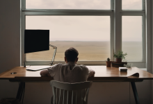 AI-generated image: A man is seated at a wooden desk in a room with a large window.