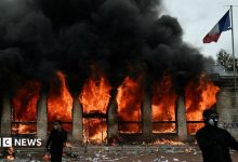 Two demonstrators, dressed all in black and in face coverings, stand outside the Lyon tourist office which is on fire behind them. A French…