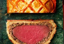 A slice of beef Wellington showing pink beef wrapped in mushroom filling and golden pastry, beside the remaining lattice-topped Wellington.