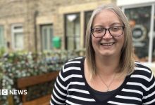 A woman with a black and white stripy top on is standing on the high street, she is wearing black rimmed glasses and has…