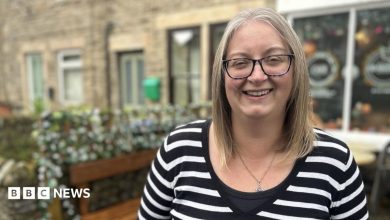 A woman with a black and white stripy top on is standing on the high street, she is wearing black rimmed glasses and has…