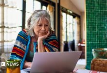 Old lady with grey hair sits at home with a brightly coloured blanket around her shoulders, looking at her laptop