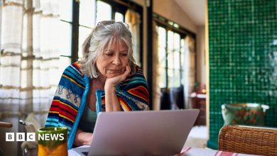 Old lady with grey hair sits at home with a brightly coloured blanket around her shoulders, looking at her laptop