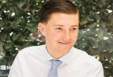 A young man smiling wearing a white shirt and a blue tie. He is sitting in front of a green bush. He has brown…