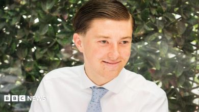 A young man smiling wearing a white shirt and a blue tie. He is sitting in front of a green bush. He has brown…