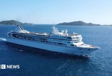 A large white cruise ship sails through deep blue ocean waters near a chain of green, hilly islands under a clear sky.