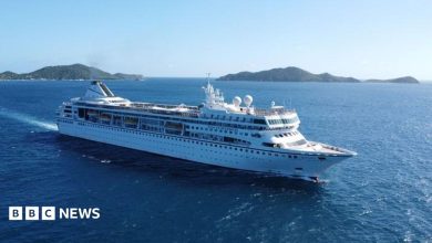 A large white cruise ship sails through deep blue ocean waters near a chain of green, hilly islands under a clear sky.