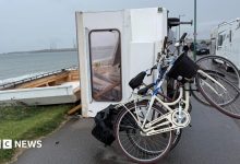 A caravan is blown in its side on a beachside road. It has two bikes attached to the back