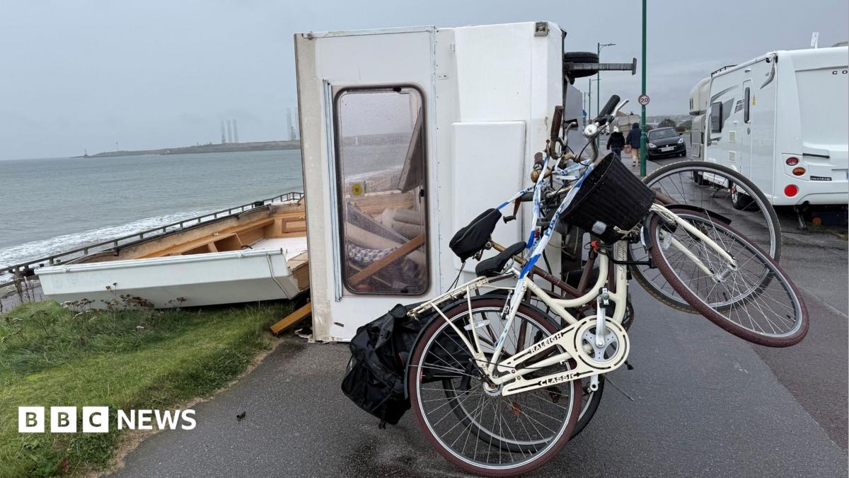 A caravan is blown in its side on a beachside road. It has two bikes attached to the back