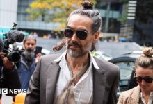Russell Brand arrives at Southwark Crown Court, alongside his wife, wearing sunglasses and with his hair tied up, as cameras fix upon him