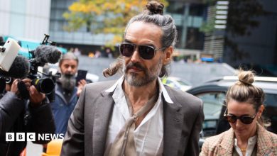 Russell Brand arrives at Southwark Crown Court, alongside his wife, wearing sunglasses and with his hair tied up, as cameras fix upon him
