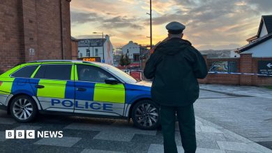 A police officer stands with his back to the camera. He is in uniform and standing beside a police car. Police tape can be…