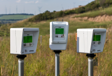 AI-generated image: Three white electronic devices with green displays are mounted on poles in a grassy field.