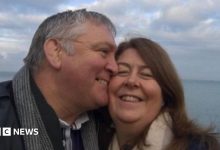 A selfie of John kissing Louisa on the cheek they are both wrapped up warm in coats and scarves and smiling as the wind…