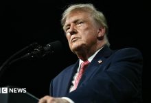 President Trump stands against a black background behing a black podium/ He is wearing a white shirt, blue suit coat with an American flag…