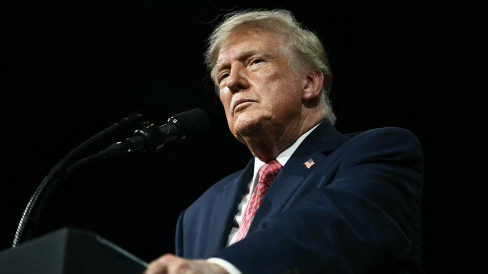 President Trump stands against a black background behing a black podium/ He is wearing a white shirt, blue suit coat with an American flag…