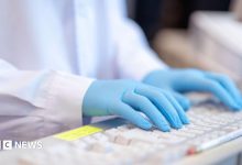 A close-up stock image of a lab worker, wearing a white coat and blue gloves, typing on a keyboard.