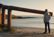 AI-generated image: A man stands on a sandy beach, wearing a grey hooded sweatshirt, a grey cap, and dark trousers with brown shoes.