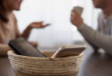Two smartphones sit in a woven basket on a table as people talk in the background, signaling a screen-free day.
