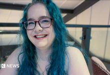 Olivia Miller-Davis, with long blue hair and glasses, sits smiling in a bar