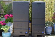 Two large black compost bins stand on a sunny garden patio, surrounded by potted pink flowers, leafy plants, and a wooden fence
