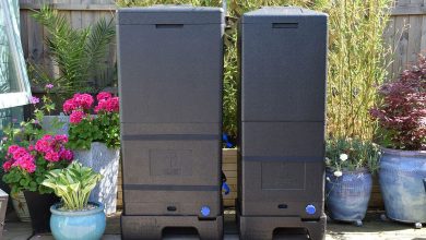 Two large black compost bins stand on a sunny garden patio, surrounded by potted pink flowers, leafy plants, and a wooden fence