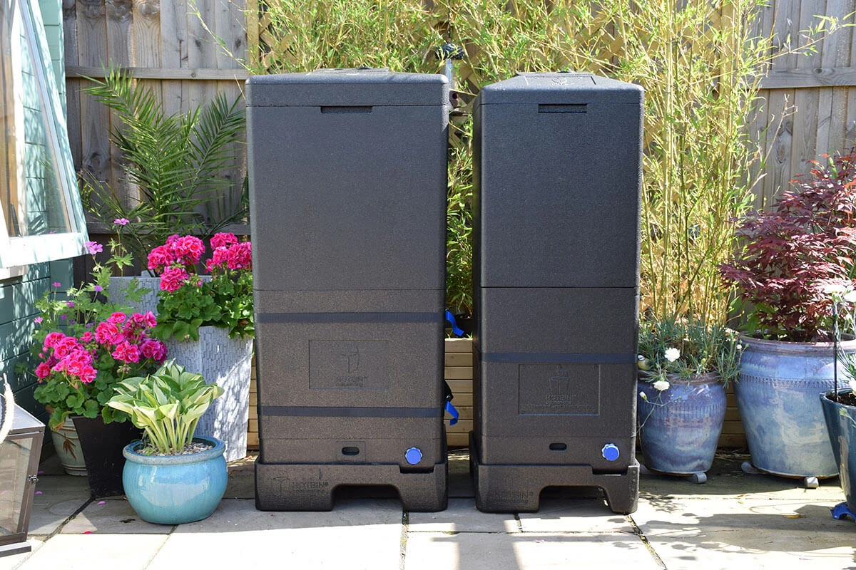 Two large black compost bins stand on a sunny garden patio, surrounded by potted pink flowers, leafy plants, and a wooden fence