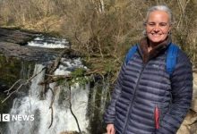 Eleanor stands by a waterfall. She has white hair, which is tied back, and wears a navy coat. She is smiling at the camera.